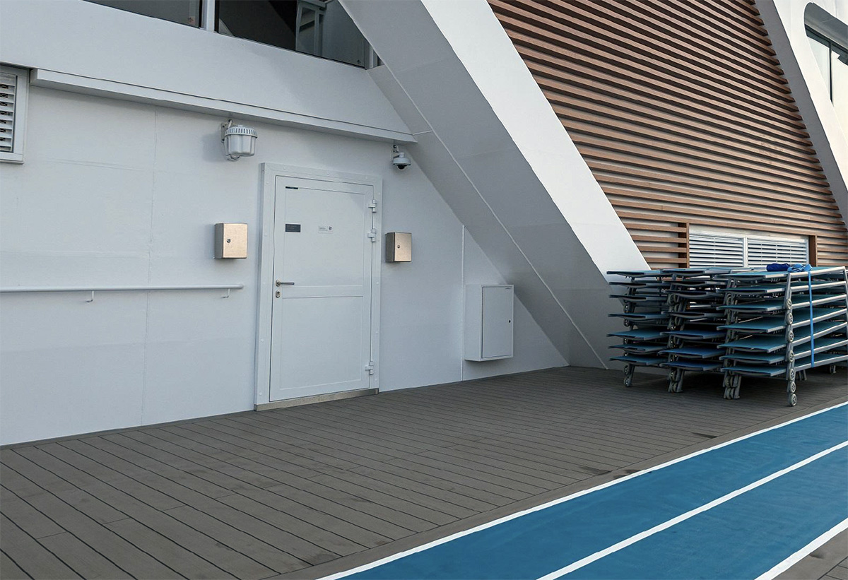 Modern exterior view on board Mein Schiff 7, featuring marine-grade aluminium-framed hinged doors designed for external use. The clean white wall contrasts with the sleek wood panel cladding and durable composite decking. The high-quality weatherproof doors are equipped with security features, suited for cruise ship environments.