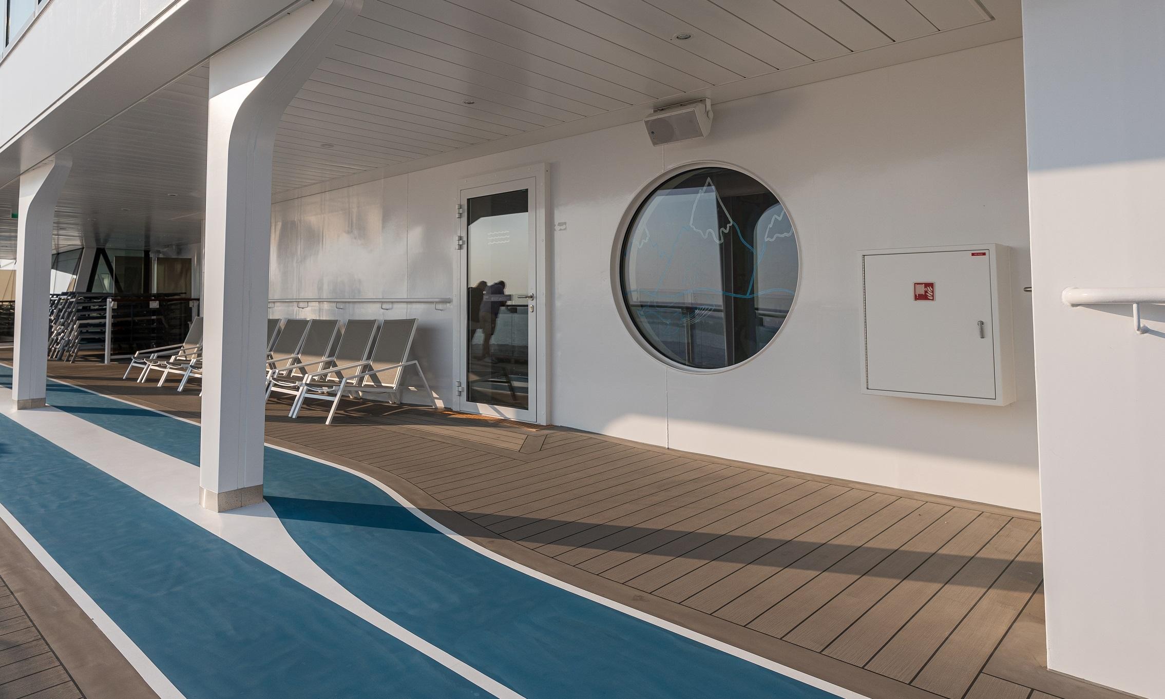 Exterior view on board Mein Schiff 7, featuring a Tarvasovi-supplied aluminum-framed hinged door with full glazing, installed along a sun deck with lounge chairs and a running track. The door is part of a larger delivery of 80 automated and manually operated doors designed for marine conditions.